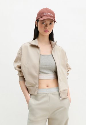 Nő, aki bézs zipzáras dzsekit, szürke sportmelltartót, hozzá illő nadrágot és rózsaszín "Marc O'Polo" sapkát visel, zsebeibe tett kézzel áll.