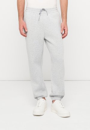 Homme portant un pantalon de survêtement gris clair avec une taille à cordon et des baskets blanches, debout devant un fond blanc uni.