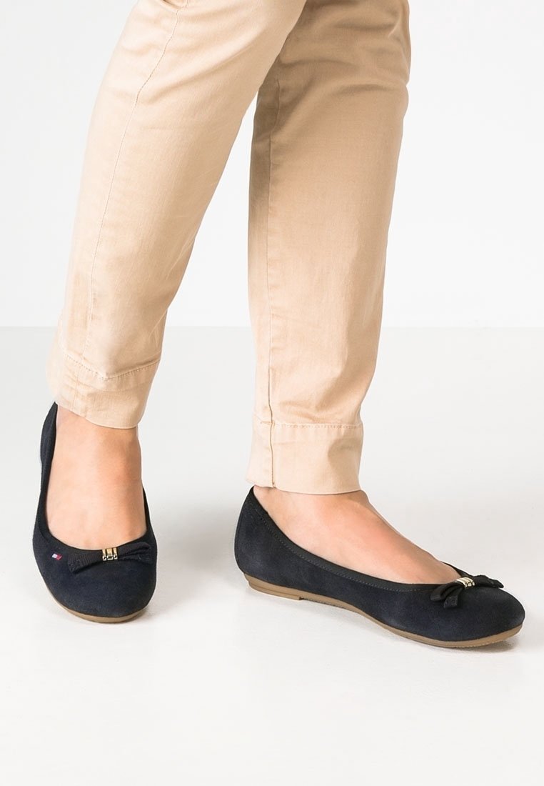 Ballerines en daim bleu marine avec un accent en nœud, bout arrondi et semelle souple et basse. Associées à un pantalon beige ajusté.