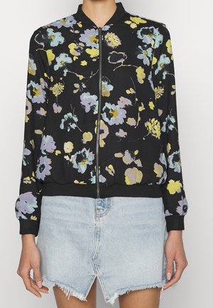 Sort lynlåstrøje med pastelgul, lilla og blå blomstermønster kombineret med lyseblå denimnederdel med frynser og frontslids.