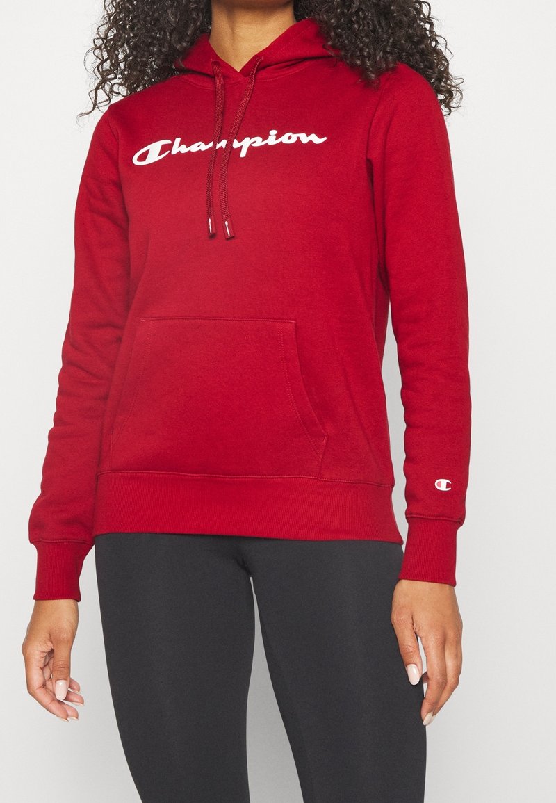 Femme portant un sweat à capuche Champion rouge avec poche avant et un legging noir, montrée de la nuque aux hanches sur un fond uni.
