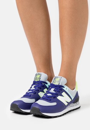 Benen met blauwe, grijze en neon groene New Balance hardloopschoenen met zwarte zolen op een witte achtergrond.