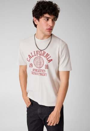 Jonge man met krullend haar draagt een wit California Athletics Department T-shirt, een ketting als halsketting, zwarte jeans en staat met één hand in zijn zak.