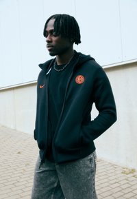 Sudadera negra con cremallera blanca, logo de Nike naranja y emblema circular naranja. Llevada sobre una camiseta negra y combinada con jeans grises.