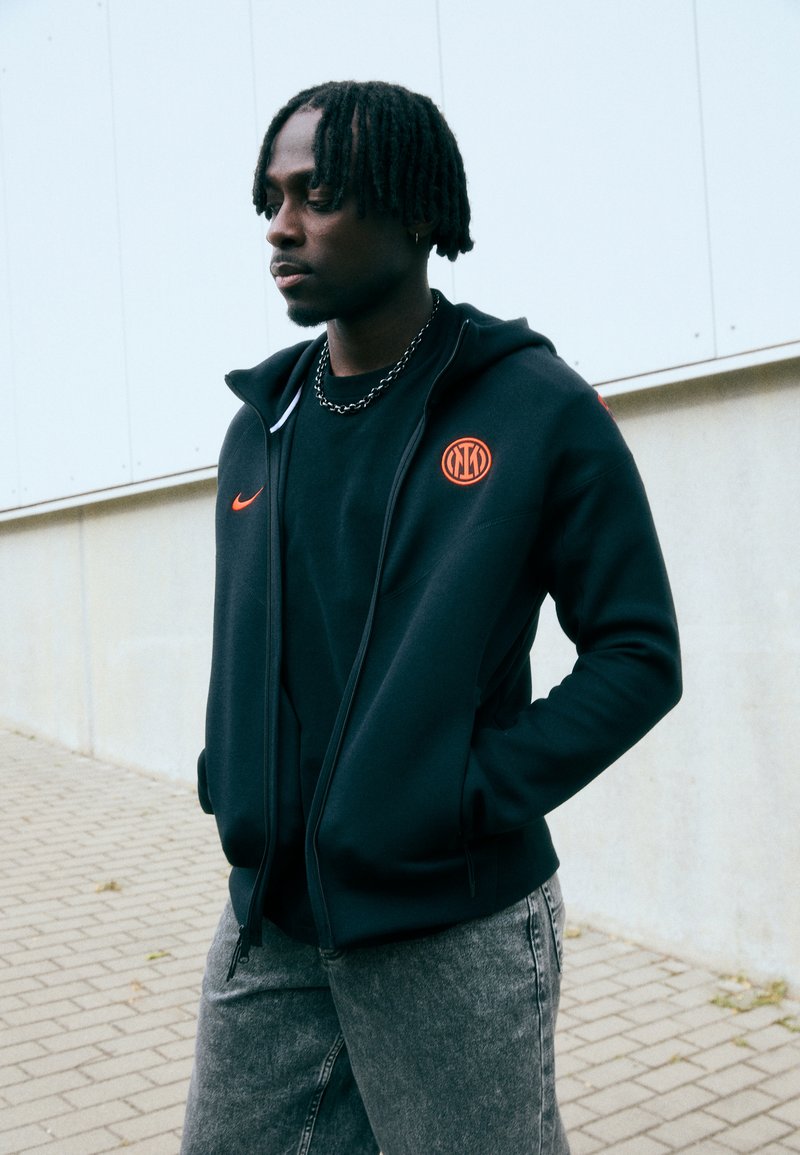 Černá zipová mikina s bílým zipem, oranžovým logem Nike a kruhovým oranžovým emblémem. Nosená přes černé tričko a spárovaná se šedými džíny.