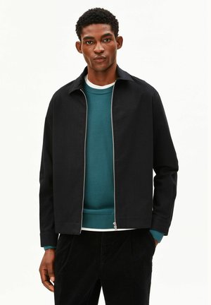 Junger Mann trägt eine schwarze Jacke mit Reißverschluss über einem türkisfarbenen Pullover und dunklen Hosen, steht mit einer Hand in der Tasche vor weißem Hintergrund.