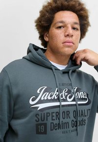 Dark green hoodie featuring a graphic print that reads "Jack & Jones Super or Quality Denim Goods" in white and black. Soft fabric texture.