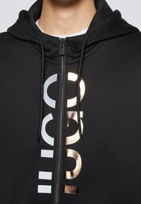 Felpa con cappuccio nera con zip, dotata di cappuccio con coulisse, scritte metalliche oro e argento sul davanti e tessuto dalla texture liscia.