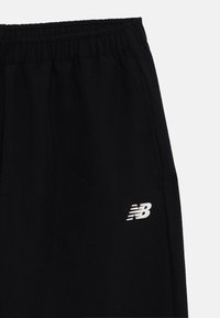 Čierne športové nohavice s elastickým pásom, bočnými vreckami a bielym logom New Balance na ľavom stehne. Mäkká, hladká textúra materiálu.