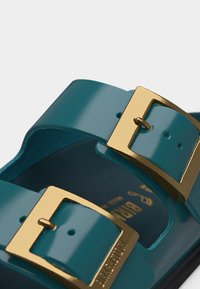 Μπλε λουριά σανδάλια με λαμπερά χρυσά ορθογώνια αγκράφες, κοντινή λήψη που δείχνει το ανάγλυφο όνομα της μάρκας στις αγκράφες και το κείμενο στη εσωτερική σόλα.