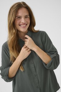 Žena sa dugom svijetlosmeđom kosom smiješi se dok zakopčava ovratnik široke tamnozelene košulje s podvrnutim rukavima.