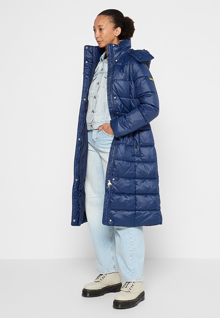 Cappotto puffer lungo navy con colletto alto, design trapuntato, chiusura con cerniera e bottoni a pressione. Indossato sopra una leggera giacca di denim e jeans, con stivali grossi.