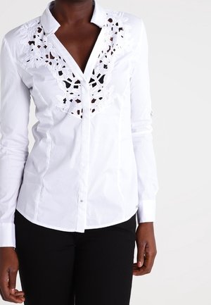 Femme portant un chemisier blanc ajusté avec des détails en dentelle florale sur le col et le devant, associé à un pantalon noir, debout devant un fond blanc.