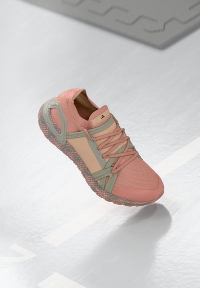 Pink and beige sneaker with green accents, textured sole, and laces, floating above a light reflective surface with a shadow beneath.