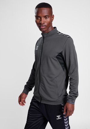 Grijze sportieve zip-up jack van textuurstof, met zwarte paneelaccenten en reflecterende details op de schouders. Gecombineerd met zwarte broek.