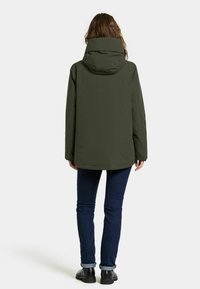 Donkergroene waterdichte jas met een hoge kraag, capuchon en een rechte snit, gecombineerd met blauwe jeans en zwarte enkellaarsjes.