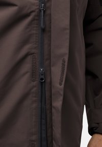 Giacca impermeabile marrone con zip frontale, dettagli in velcro e una texture liscia. Dispone di tasche laterali e un design snello.