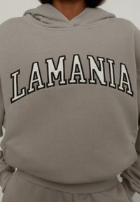 Hanorac gri cu cuvântul "LAMANIA" scris cu litere îndrăznețe în alb și negru. Materialul are o textură moale și manșete și tiv nervurate.