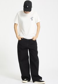 Hvid bomulds t-shirt med et lille blåt broderet design, parret med løse sorte denim bukser og sorte sneakers med en cremefarvet sål.
