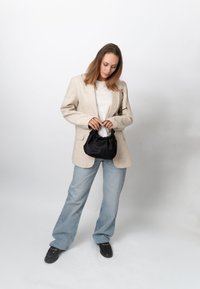 Frau trägt beige Blazer und hellblaue Jeans, hält eine kleine schwarze Handtasche und blickt auf diese hinunter vor einem einfarbig weißen Hintergrund.