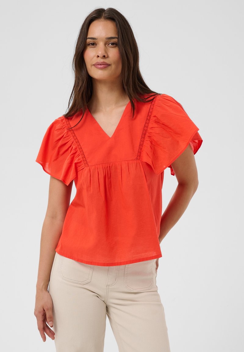 Femme aux longs cheveux foncés, portant un chemisier orange vif à manches courtes avec des épaules volantées et un pantalon beige sur un fond uni.