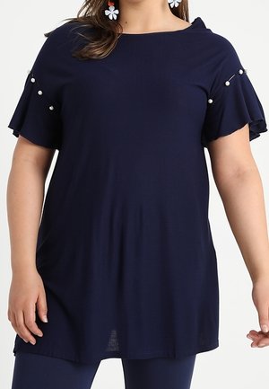 Frau trägt eine marineblaue Tunika mit kurzen, gerafften Ärmeln, die mit Perlen verziert sind, kombiniert mit passenden marineblauen Leggings und weißen Blumenohrringen.