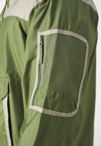 Chaqueta verde con un bolsillo con cremallera y un panel de malla en la manga, acentuada con un ribete beige a lo largo del borde.
