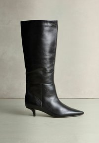 Fekete bőr, térd feletti csizma, sima hegyes orral és alacsony, keskeny sarokkal. Jellemzi a sima textúra és a minimalista dizájn.