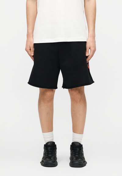 Άτομο που φοράει μαύρο σορτς, λευκές κάλτσες, μαύρα αθλητικά παπούτσια και λευκό πουκάμισο, στέκεται μπροστά σε ένα απλό φωτεινό φόντο.