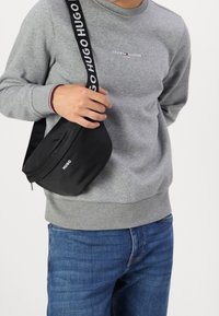 Schwarze Nylon-Crossbody-Tasche mit "HUGO"-Logo, geschwungener Form und verstellbarem Schultergurt, getragen mit einem grauen Sweatshirt und blauen Jeans.