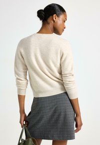 Donna con capelli scuri raccolti in un bun, indossa un maglione color crema e una gonna a quadretti, tiene una borsa verde, rivolta di lato su uno sfondo bianco.