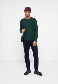 Grüner Strickpullover, marineblaue Hose, schwarze Slipper und eine kastanienbraune Mütze. Das Model hält eine schwarze Umhängetasche mit einem Riemen. Einfaches, körperbetontes Design.