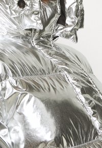 Skimrande silverfärgad puffjacka med hög krage, vadderad textur, synlig sömnad och tryckknappsdetaljer.