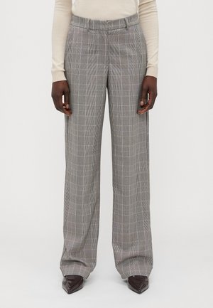 Persoon draagt grijze geruite broek met wijde pijpen en voorzakken, gecombineerd met een beige top met lange mouwen en donkerbruine puntige schoenen.