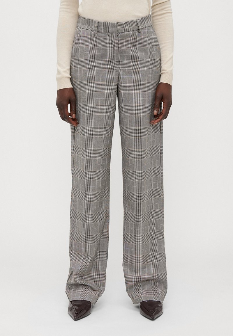 Personne portant un pantalon large à carreaux gris avec poches avant, associé à un haut beige à manches longues et des chaussures pointues marron foncé.