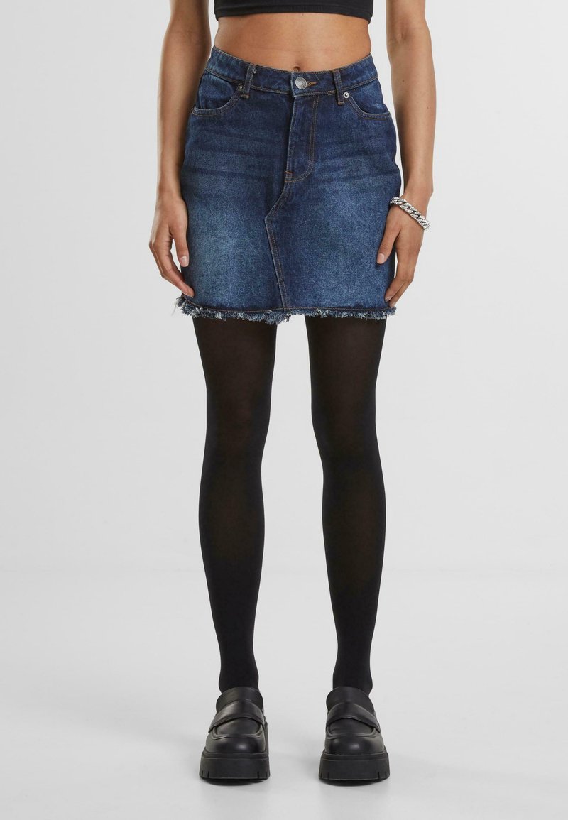 Denim mini-rok met een rafelige zoom, met een hoge taille, voorkwabben en een blauwe wasservice. Geddragen met zwarte ondoorzichtige panty’s en stevige zwarte schoenen.