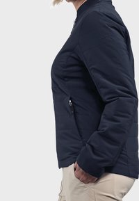 Granatowa pikowana kurtka z wysokim kołnierzem, bocznymi kieszeniami na zamek oraz minimalistycznym logo na rękawie. Materiał wydaje się gładki i lekki.
