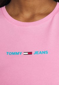 Růžové bavlněné tričko s vyšitými nápisy "TOMMY" a "JEANS" v tyrkysové barvě, s obdélníkovým logem v červené, bílé a černé.