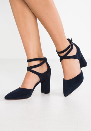 Granatowe zamszowe buty na wysokim obcasie z noskiem w szpic, paskami wokół kostki i szerokim obcasem. Charakteryzują się gładką teksturą i eleganckim wzornictwem.