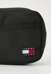 Svart nylonpung med en teksturert overflate, som har en flerfarget "TOMMY JEANS" logo-lapp og en glidelås-lukking.