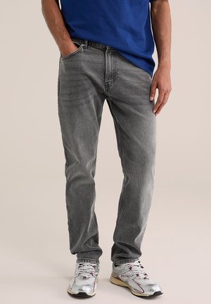 Uomo con jeans grigi sbiaditi, scarpe da ginnastica bianche e una camicia blu, in piedi con una mano in tasca su sfondo semplice.