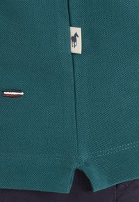Tissu texturé en teal avec un petit emblème de drapeau cousu et une étiquette blanche présentant la silhouette d'un cheval marine près d'une couture.