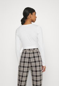Camisola branca de mangas longas com um detalhe amarrado nas costas, combinada com calças de cintura alta em padrão xadrez nas tonalidades preta, creme e castanho.