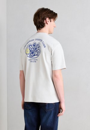 Νεαρός άνδρας που φοράει λευκό μπλουζάκι με μπλε γραφικό κοχυλιού στρειδιού και λεμονιού και το κείμενο "Lindbergh Oyster Club Sunday Nights 5PM-8PM" στην πλάτη.