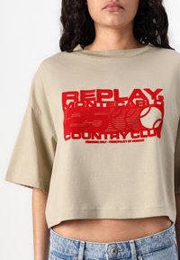 Beige cropped t-shirt med rund halsudskæring og korte ærmer, der har fed rød tekst og tennistema-designelementer i midten.
