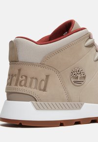 Beige Wildlederschuh mit rotem Stoffkragen, weißer Gummisohle, eingeprägtem Logo und strukturierten Akzenten. Schnürverschluss für einen sicheren Sitz.