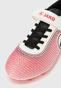 Κόκκινο-λευκό παπούτσι ποδοσφαίρου με ζιγκ-ζαγκ μοτίβο, μαύρα κορδόνια και ιμάντα με βέλκρο. Διαθέτει λογότυπο στη πλευρά και ανάγλυφη επάνω επιφάνεια.