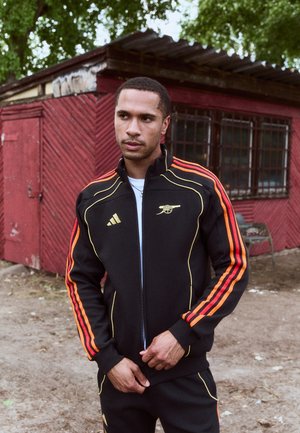 Zwarte zip-up trainingsjack met oranje en gele strepen, voorzien van een geborduurd logo. De stof is glad, met een aansluitend ontwerp.