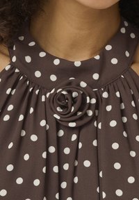 Robe marron à pois blancs, avec un col rond et un détail en forme de rose en tissu au centre devant.