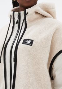 Cremefärgad fleecejacka med kontrasterande svart dragkedja, huva och detaljer; har en liten svart etikett med New Balance-logotypen.